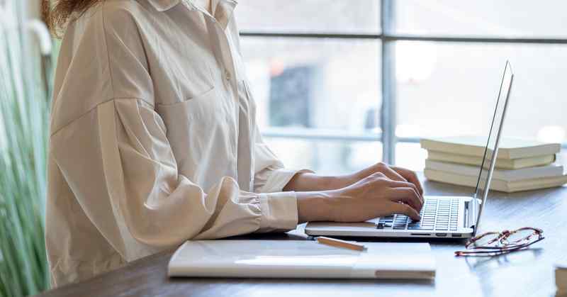 A woman at a desk, typing at a laptop, with some documents, a pen, and a pair of glasses on the desk