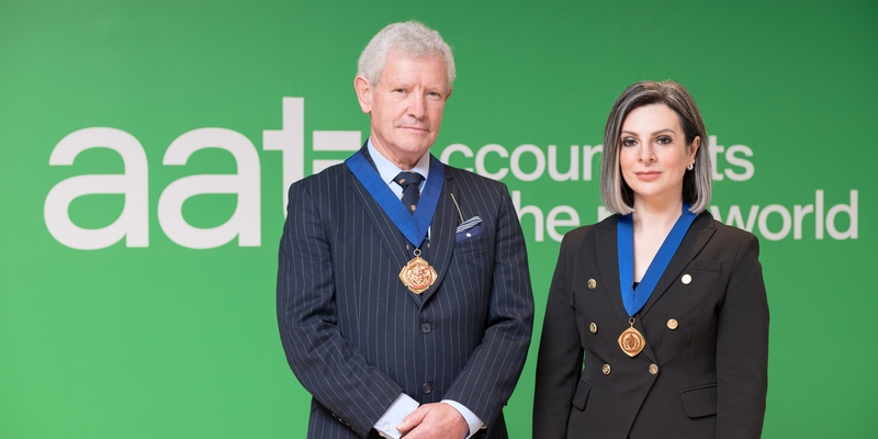 AAT President Michael Steed and AAT Vice-President Lucy Cohen standing in front of AAT corporate logo and surrounding green wall. They have medals on.